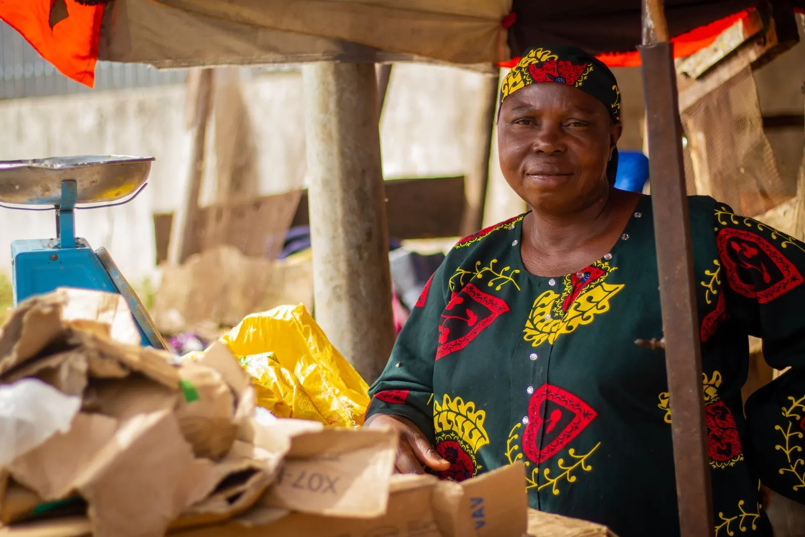 A smiling woman at her outdoor market stall, with a weighing scale beside her.